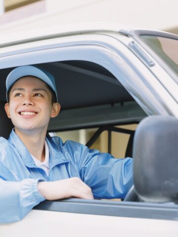 A person wearing a light blue cap and jacket is sitting in the driver's seat of a white vehicle, smiling and looking out the window, reflecting the importance of Uber driver safety.