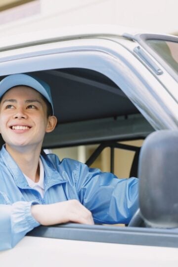 A person wearing a light blue cap and jacket is sitting in the driver’s seat of a white vehicle, smiling and looking out the window, reflecting the importance of Uber driver safety.