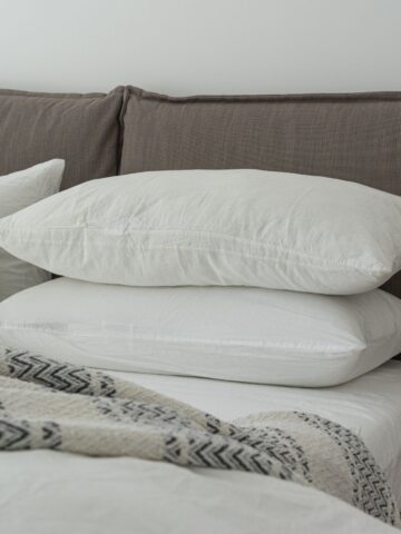 Two white pillows stacked on a bed with white sheets, a beige and gray patterned blanket, and a brown upholstered headboard-perfect for relaxing after following a simple pillow cleaning or washing guide.