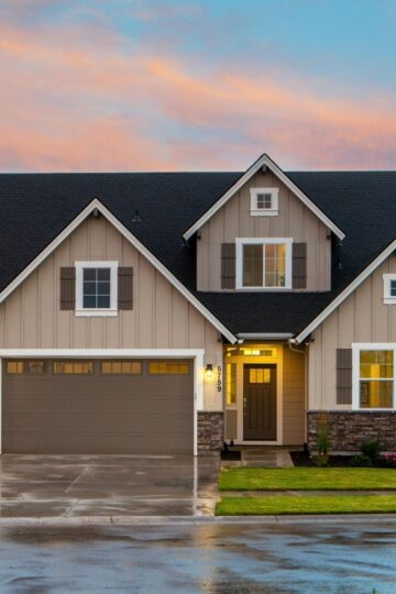 A modern suburban house with three garage doors, stone and siding exterior, front lawn, and lights turned on at sunset—designed for home safety and comfort, supporting aging in place for seniors.