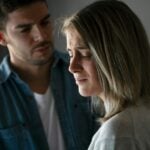 A woman with a distressed expression looks down while a man stands behind her, speaking with a serious look, reflecting on how early experiences can shape adult relationships.