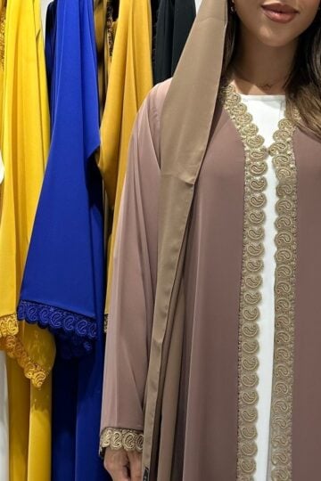 A woman wearing a brown abaya from an abaya studio stands beside a rack of Crystal Abayas in white, yellow, blue, and mustard, each with decorative trim—offering elegant options for modest clothing.