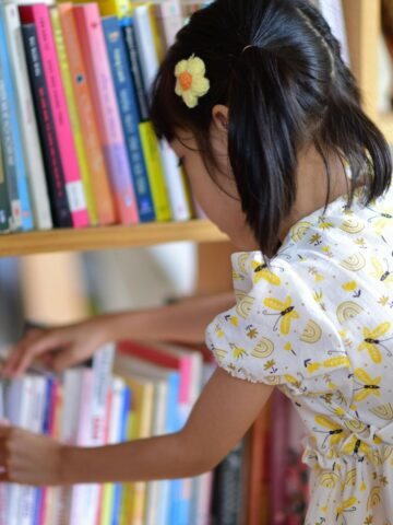 A young girl in a patterned dress selects a book from a shelf filled with colorful books, embracing accessible learning and fostering school literacy.