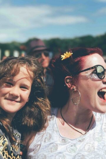 A smiling child and an excited adult wearing sunglasses and a flower hair accessory stand together outdoors at a crowded culture festival under a blue sky.
