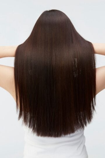 A person with straight, long, dark hair—reflecting great hair health—is shown from the back, wearing a white sleeveless top and holding their hands behind their head.