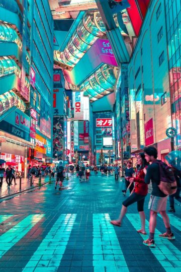 People walk across a brightly lit street in a busy city area at night, surrounded by tall buildings and colorful neon signs—an exciting scene for those who want to explore Tokyo and enjoy stress-free travel.