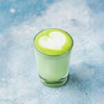 A glass of green matcha latte with a heart-shaped design in the foam, placed on a light blue textured surface-a perfect example of beverage trends transforming the beverage industry.