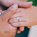 A close-up of two hands holding, one wearing the perfect engagement ring-a large oval diamond-and a matching wedding band with small diamonds.
