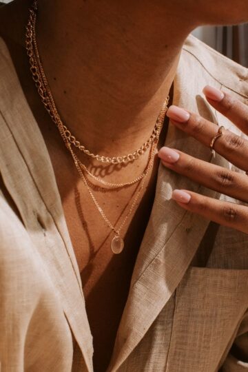 A person wearing real jewelry—layered gold necklaces and rings—with neatly manicured nails, dressed in a light tan linen shirt.