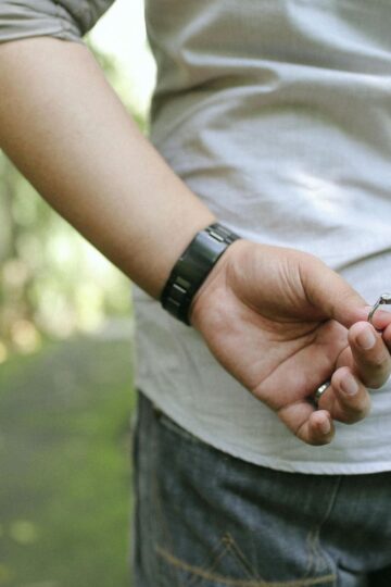 A person holds a ring behind their back, standing outdoors with another person blurred in the background—capturing the anticipation of a marriage proposal.