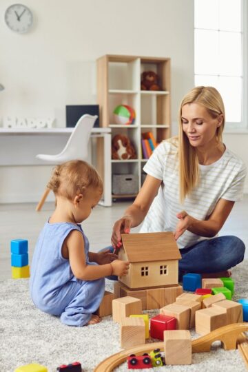 An adult and a toddler sit on a carpet in a mom-friendly home, playing with wooden blocks and a toy house, surrounded by colorful toys in a thoughtfully organized living room.