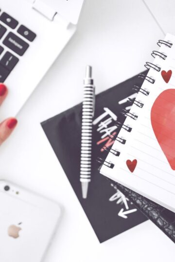 A person with red nail polish uses a laptop next to a spiral notebook with a large red heart—perhaps searching for finding your perfect match online—plus a striped pen, black card, and iPhone.