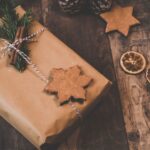 A brown paper-wrapped gift decorated with a pine sprig, cinnamon sticks, and a star-shaped cookie on twine-perfect for seasonal gift ideas. Pine cones, dried citrus slices, and another cookie sit nearby for festive inspiration.