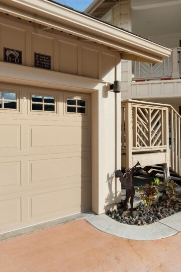 Beige garage door and staircase leading to a porch, with a metal horse sculpture and landscaped plants along the edge of the driveway.