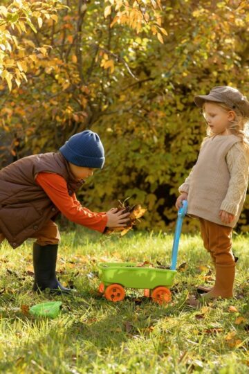Two children gather autumn leaves in a sunny yard; one collects leaves while the other holds a small green wagon, dreaming of starting an edible garden and discovering the joys of gardening with kids.