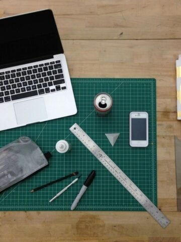 A laptop, smartphone, ruler, pens, pencil, cutting mat, soda can, notebook, and various stationery items arranged on a wooden desk reflect the blend of technology and the importance of art in today's tech-driven world.