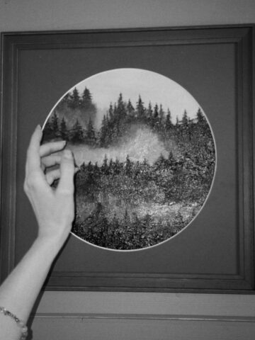 A hand reaches toward a framed circular print of a misty forest, reproduced with precision scanning, hung on a paneled wall.