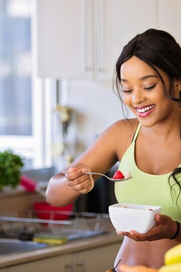 Woman standing in a kitchen, smiling with self-confidence while eating from a white bowl with a spoon—embracing the joy of living well.