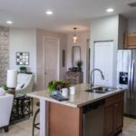 Modern kitchen with granite island, stainless steel appliances, and limestone floors. White armchairs and decorative accents in the living area offer kitchen design ideas to elevate your space.