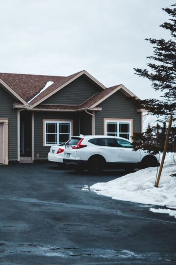 A white SUV is parked in the big driveway of a gray house with a two-car garage; patches of snow linger on the ground, perfect for ticking off your Winter home prep checklist, while a tree stands near the driveway.