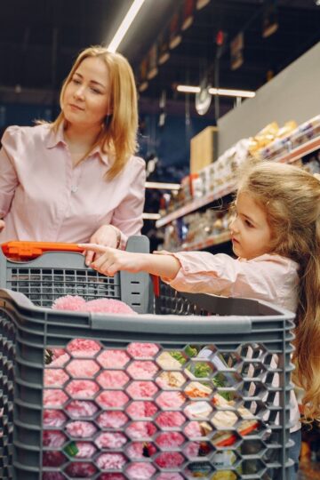 An adult and a child with long hair are shopping in a grocery store, carefully pushing a cart filled with various items down an aisle to avoid a store slip.