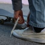 A person in jeans ties the laces of their white sneakers next to a skateboard on a paved surface, ready to start their next generation journey.