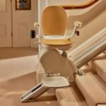 A Stannah stairlift with a tan seat and footrest is installed on a carpeted staircase in a residential home.