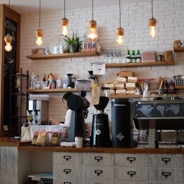 Modern coffee shop interior with wooden counter, hanging lights, espresso machines, shelves with cups and supplies, and a chalkboard menu on the wall.