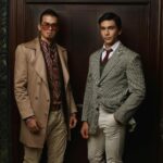 Two men stand indoors in front of a wooden door, both wearing stylish, vintage-inspired outfits with patterned shirts and blazers, showcasing how the right fabric can elevate a personalized men's suit.