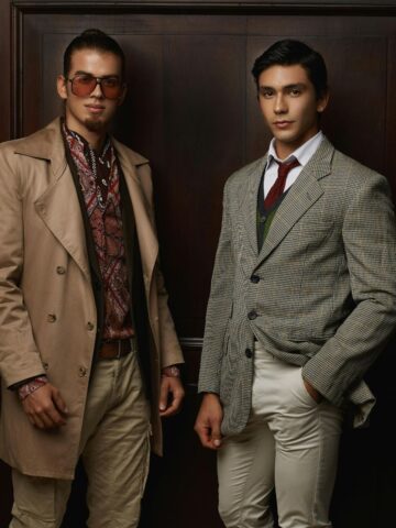 Two men stand indoors in front of a wooden door, both wearing stylish, vintage-inspired outfits with patterned shirts and blazers, showcasing how the right fabric can elevate a personalized men's suit.