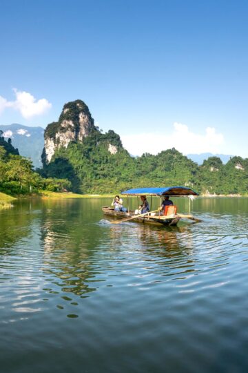 A small boat with people on a calm lake, surrounded by green hills and rocky mountains under a clear blue sky, offers a peaceful slow travel experience immersed in nature.