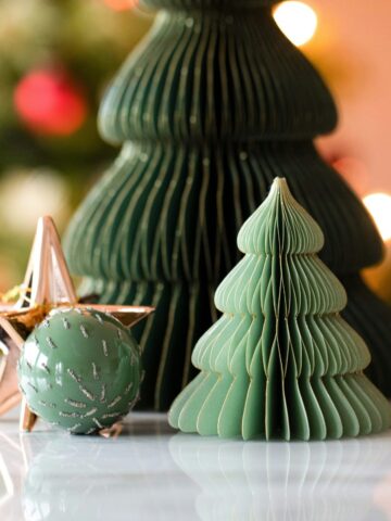 Two green paper Christmas tree decorations sit on a reflective surface, next to a green ornament with silver glitter and a metallic star. Blurred Christmas lights add festive cheer, perfect for busy families in Dallas preparing for the holidays.