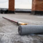 A paint roller lies on a concrete surface with its handle extended in the background, near brick columns-hinting at ongoing garage transformation.