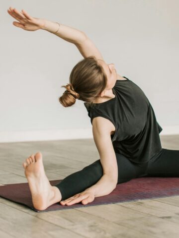 Person on a yoga mat performing a seated side stretch, with legs extended and one arm reaching overhead in a bright indoor space-an ideal pose for improving flexibility and supporting healthy ageing.