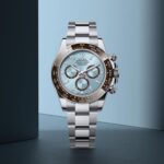 A Rolex wristwatch with a silver metal band, brown bezel, and light blue face with three subdials, displayed upright against a blue background.