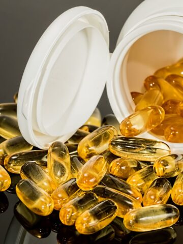 An open white bottle with yellow gel capsules, rich in Omega-3s for muscle recovery, spills out onto a reflective black surface.