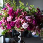 Large ornate silver vase filled with an arrangement of pink, magenta, and purple flowers with green foliage, set on a wooden table indoors.