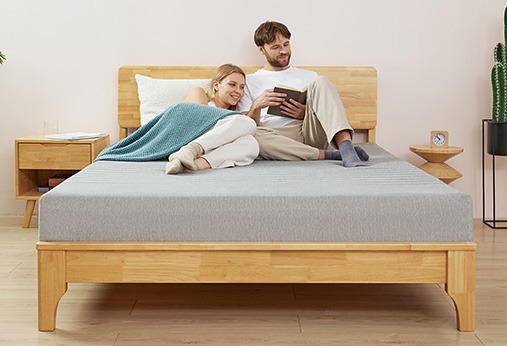 A couple sits on a wooden bed with a gray mattress; one person is reading a tablet while the other leans on them. Nearby are a nightstand, small table, and houseplant.