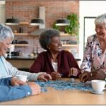 Five older adults sit around a table working on a jigsaw puzzle and talking, with coffee cups nearby in a kitchen setting.