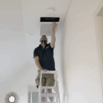 A man standing on a ladder inspects or adjusts a ceiling vent in a bright room, showcasing the clean look of ducted air conditioning-the best choice for modern interiors.
