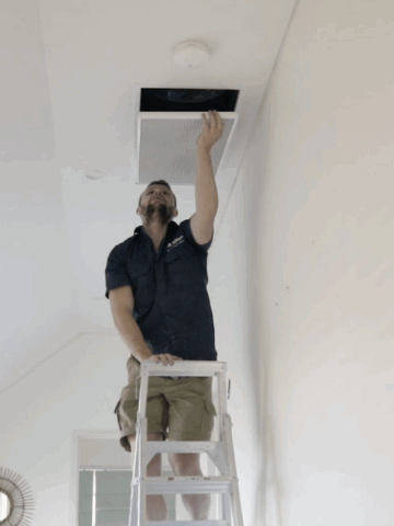 A man standing on a ladder inspects or adjusts a ceiling vent in a bright room, showcasing the clean look of ducted air conditioning-the best choice for modern interiors.
