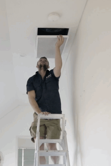 A man standing on a ladder inspects or adjusts a ceiling vent in a bright room, showcasing the clean look of ducted air conditioning—the best choice for modern interiors.
