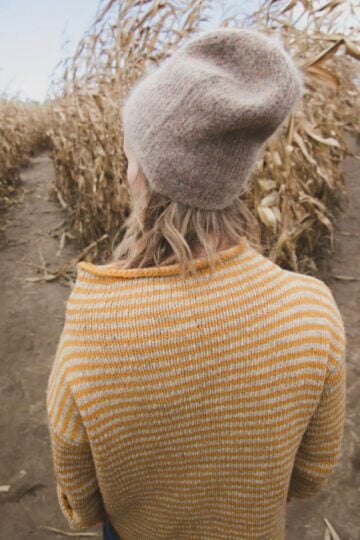 Person in a striped sweater and knit hat stands facing a fork in a cornfield path, surrounded by tall dried cornstalks—capturing a quiet moment of decision-making amid nature.