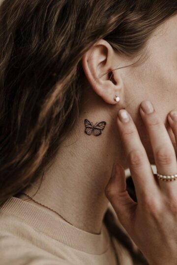 A woman with wavy brown hair shows a small black butterfly tattoo behind her ear. She wears a beige top, stylish flat-back earrings, a pearl earring, and rings—perfect accessories for chic dinner dates.