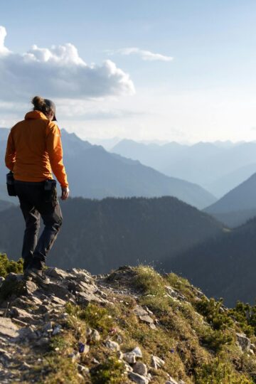 Person wearing an orange jacket stands on a rocky mountain ridge, looking out over layers of distant, forested mountains under a partly cloudy sky—perfect for discovering travel ideas for introverts seeking serene adventures.