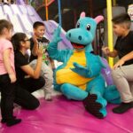 Four children sit and stand around a person in a blue dragon costume, giving high fives on a colorful indoor playground-perfect for indoor family fun and year-round activities in Tampa.