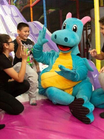 Four children sit and stand around a person in a blue dragon costume, giving high fives on a colorful indoor playground-perfect for indoor family fun and year-round activities in Tampa.