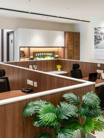 Modern office workspace with wooden cubicles, black chairs, computers, potted plants, and wall cabinets, featuring a large poster on the wall and bright, even lighting to boost productivity.