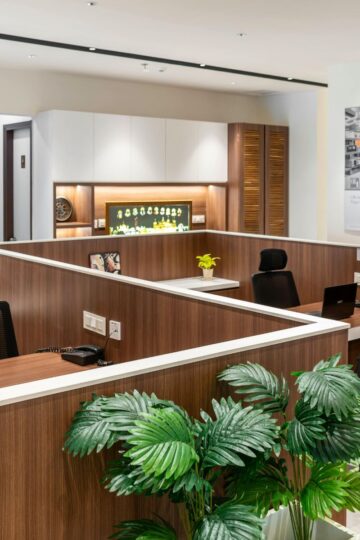 Modern office workspace with wooden cubicles, black chairs, computers, potted plants, and wall cabinets, featuring a large poster on the wall and bright, even lighting to boost productivity.