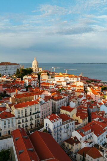 Aerial view of Lisbon’s Alfama district showing red-roofed buildings, a church dome, and the Tagus River under a partly cloudy sky.